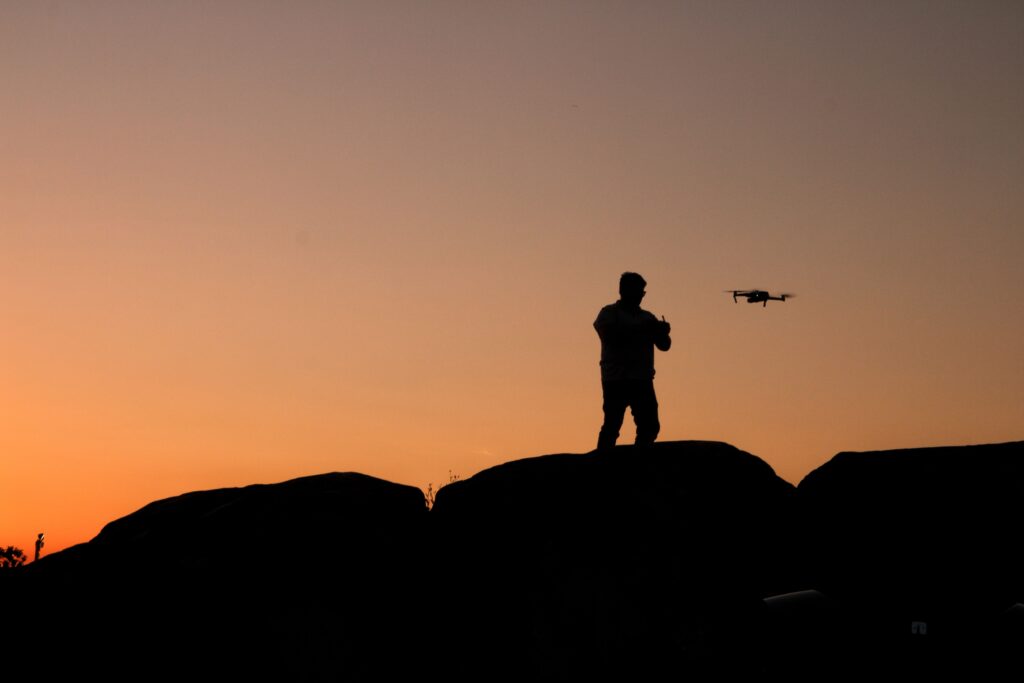 Silhouette of a man controlling a drone at sunset with rocky foreground.